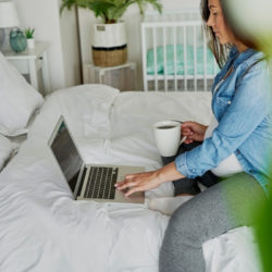 Schwangere Frau sitzt mit Laptop und Kaffee auf dem Bett